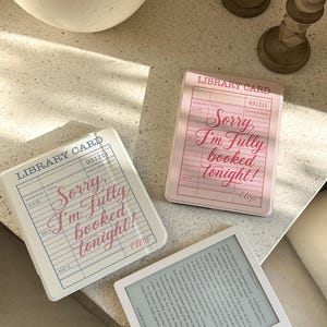 May include: Three coasters with a library card design and the text "Sorry, I'm Fully booked tonight!" in pink and blue. A white e-reader with text is also present. The coasters are in shades of pink and white.