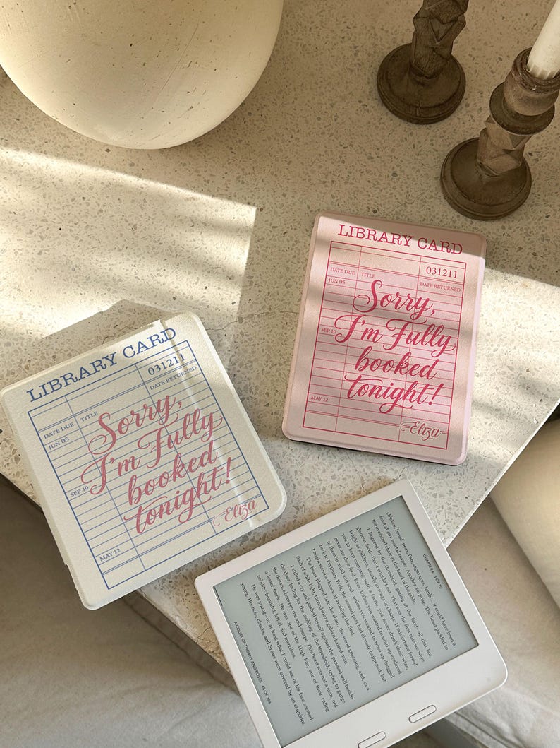 May include: Three coasters with a library card design and the text "Sorry, I'm Fully booked tonight!" in pink and blue. A white e-reader with text is also present. The coasters are in shades of pink and white.