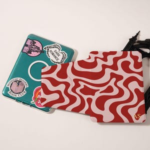 Puede incluir: Una funda para port&aacute;til de color turquesa con pegatinas y un tarjetero con un patr&oacute;n ondulado rojo y rosa. Las pegatinas incluyen "Dark Romance" y "Midnight Readers Book Club". El tarjetero tiene las iniciales "AS".