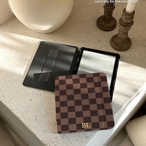 May include: A black e-reader case with a brown and dark brown checkerboard pattern on the front cover. The cover features the gold foil initials BL at the bottom. The interior is black with a hand strap. Text reads: Kobo Libra Colour 7" / Kobo Libra 2 7".