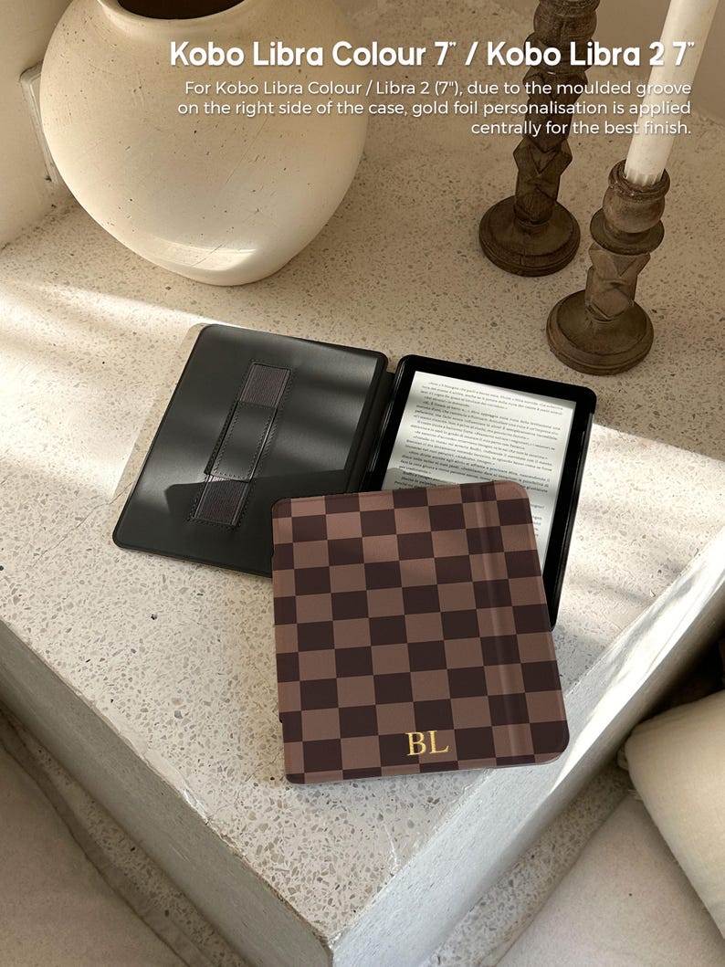 May include: A black e-reader case with a brown and dark brown checkerboard pattern on the front cover. The cover features the gold foil initials BL at the bottom. The interior is black with a hand strap. Text reads: Kobo Libra Colour 7" / Kobo Libra 2 7".