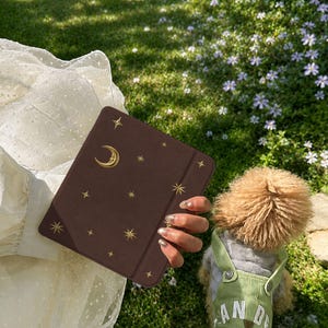 May include: A dark brown rectangular e-reader case featuring a gold-colored crescent moon and star pattern. The case has a smooth, matte finish and a vertical elastic band on the right side for closure, designed to protect a digital tablet device.