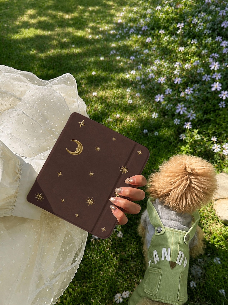 May include: A dark brown rectangular e-reader case featuring a gold-colored crescent moon and star pattern. The case has a smooth, matte finish and a vertical elastic band on the right side for closure, designed to protect a digital tablet device.