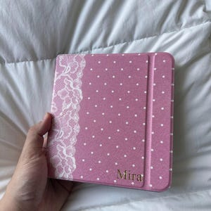 May include: A pink tablet case featuring a white polka dot pattern and a vertical strip of white lace detailing along the left side. The name Mira is printed in gold lettering in the bottom right corner of the textured, rectangular cover.