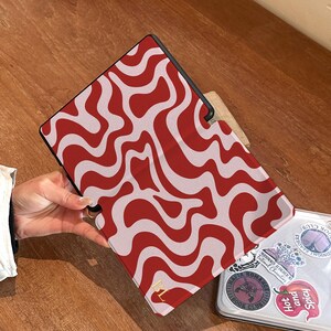Puede incluir: Funda para tableta con un dise&ntilde;o ondulado rojo y blanco. La funda est&aacute; abierta, revelando un port&aacute;til con pegatinas. Un logotipo dorado es visible en la funda. El fondo es una superficie de madera.