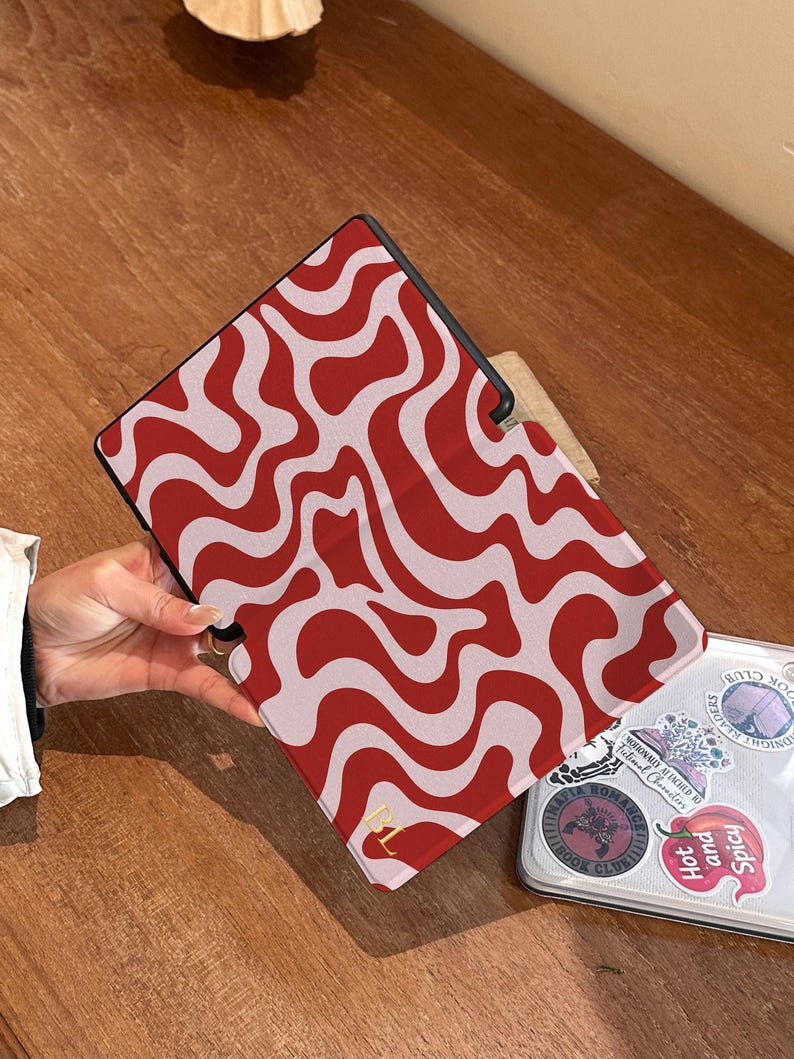 Puede incluir: Funda para tableta con un dise&ntilde;o ondulado rojo y blanco. La funda est&aacute; abierta, revelando un port&aacute;til con pegatinas. Un logotipo dorado es visible en la funda. El fondo es una superficie de madera.