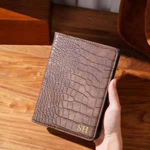 May include: A brown, textured wallet with a crocodile skin pattern. The wallet has the gold initials "SH" on the lower right corner. The wallet is held by a person's hand.