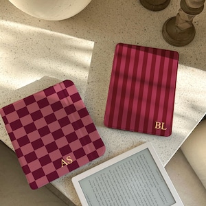 Könnte beinhalten: Drei rosa und rot gemusterte Hüllen, zwei mit Initialen, liegen auf einer hellen Oberfläche neben einem E-Reader. Eine Hülle hat ein Schachbrettmuster, eine andere vertikale Streifen, und die dritte ist einfarbig. Der E-Reader zeigt Text an.