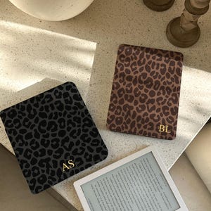 Könnte beinhalten: Drei E-Reader-Hüllen mit Leopardenmuster in Grau, Braun und Beige, jeweils mit goldenen Initialen. Ein weißer E-Reader zeigt Text an. Die Hüllen liegen auf einer hellen Oberfläche.