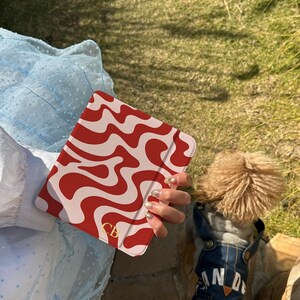 Puede incluir: Una funda para tableta con estampado rojo y blanco con las iniciales doradas "CB". La funda se sostiene frente a un vestido azul claro y un perro con un peto vaquero. El peto del perro tiene la palabra "AND".