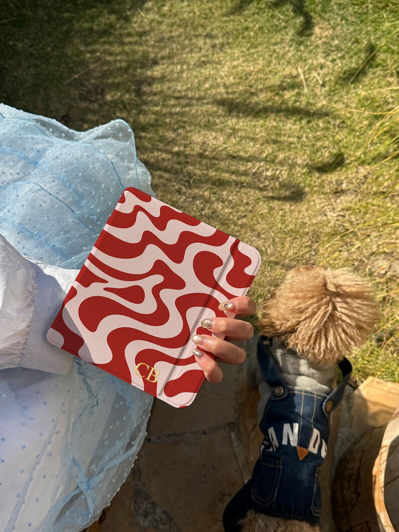 Puede incluir: Una funda para tableta con estampado rojo y blanco con las iniciales doradas "CB". La funda se sostiene frente a un vestido azul claro y un perro con un peto vaquero. El peto del perro tiene la palabra "AND".
