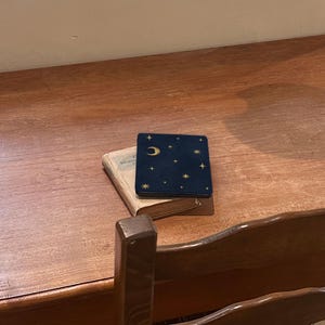 May include: A dark blue square case with a gold crescent moon and star pattern resting on top of a small, weathered, light-colored hardcover book. The case features a smooth, matte finish with celestial gold foil accents.