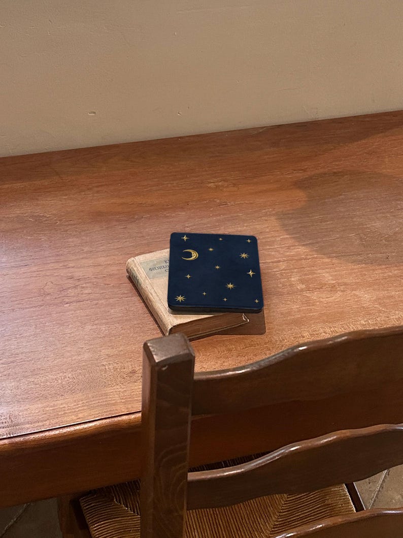 May include: A dark blue square case with a gold crescent moon and star pattern resting on top of a small, weathered, light-colored hardcover book. The case features a smooth, matte finish with celestial gold foil accents.