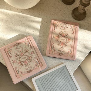 Puede incluir: Tres fundas rosas para lectores electrónicos con diseños florales y de mariposas. Las fundas presentan el nombre "Eliza". Una funda está abierta, revelando la pantalla del lector electrónico con texto. Las fundas están sobre una superficie clara.