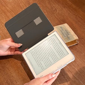 May include: A white Kobo e-reader with a clear protective case and a black folding cover featuring a gray elastic hand strap. The screen displays text from a book, and the device is held open, revealing the digital interface and the physical cover.