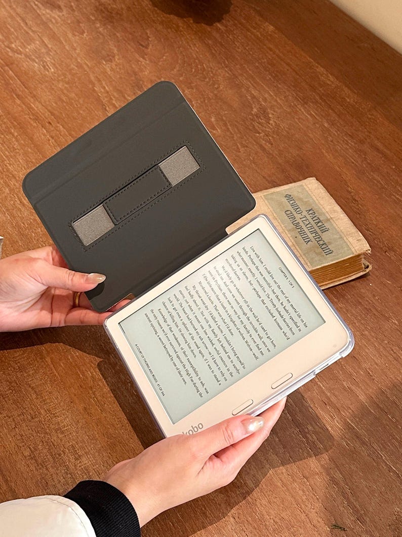 May include: A white Kobo e-reader with a clear protective case and a black folding cover featuring a gray elastic hand strap. The screen displays text from a book, and the device is held open, revealing the digital interface and the physical cover.