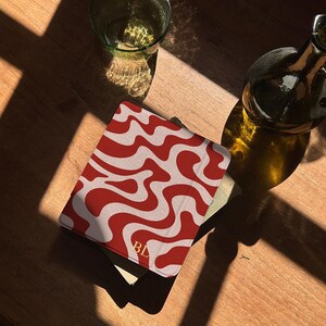 Puede incluir: Una cubierta de libro con estampado rojo y blanco con las iniciales "BL" en dorado. La cubierta est&aacute; sobre una superficie de madera, junto a un vaso y una botella. El dise&ntilde;o presenta un patr&oacute;n ondulado.