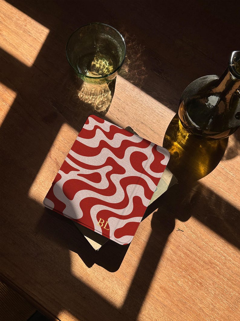 Puede incluir: Una cubierta de libro con estampado rojo y blanco con las iniciales "BL" en dorado. La cubierta est&aacute; sobre una superficie de madera, junto a un vaso y una botella. El dise&ntilde;o presenta un patr&oacute;n ondulado.