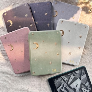 May include: A collection of six rectangular e-reader cases in various colors including dark gray, purple, light gray, pink, green, and white, each featuring gold foil crescent moon and star patterns. Two cases display gold monogrammed initials.