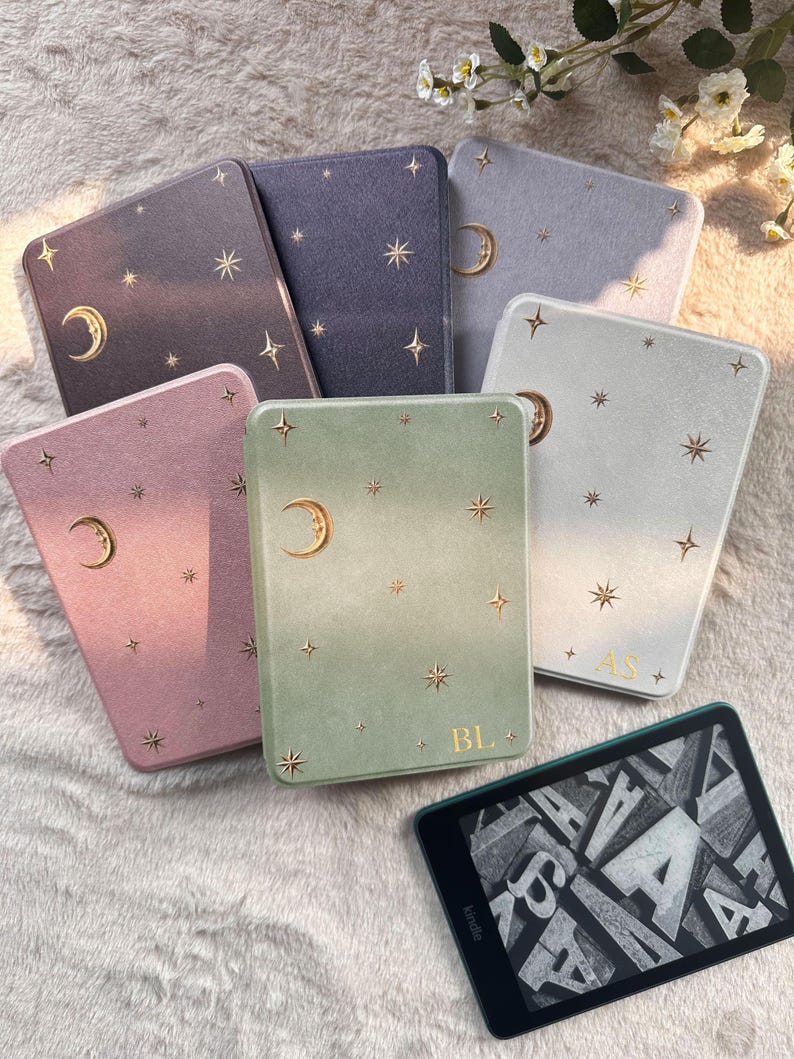 May include: A collection of six rectangular e-reader cases in various colors including dark gray, purple, light gray, pink, green, and white, each featuring gold foil crescent moon and star patterns. Two cases display gold monogrammed initials.