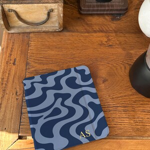Puede incluir: Una funda para tableta azul marino con un patr&oacute;n ondulado azul claro. Las iniciales doradas "AS" est&aacute;n en la esquina inferior derecha. La funda est&aacute; sobre una superficie de madera.