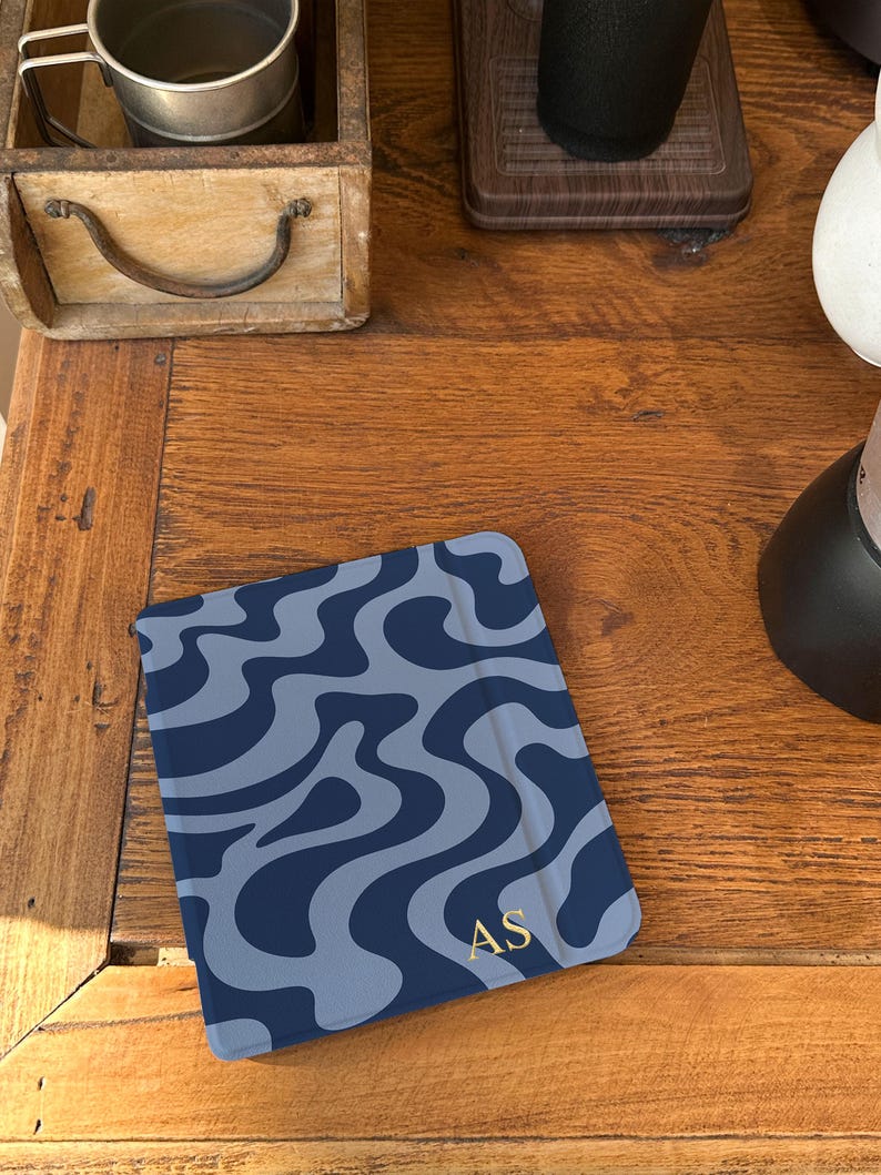 Puede incluir: Una funda para tableta azul marino con un patr&oacute;n ondulado azul claro. Las iniciales doradas "AS" est&aacute;n en la esquina inferior derecha. La funda est&aacute; sobre una superficie de madera.
