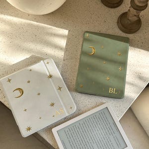 May include: A white e-reader case with gold crescent moon and star accents and the initials AS, a sage green e-reader case with gold moon and star accents and the letters BL, and a white e-reader displaying text from A Court of Thorns and Roses.