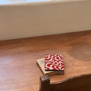 Puede incluir: Un posavasos con un patr&oacute;n ondulado rojo y blanco descansa sobre un libro peque&ntilde;o en una mesa de madera. El dise&ntilde;o del posavasos es un patr&oacute;n repetitivo de l&iacute;neas onduladas. Una silla de madera y una ventana con un vaso verde y un jarr&oacute;n est&aacute;n en el fondo.