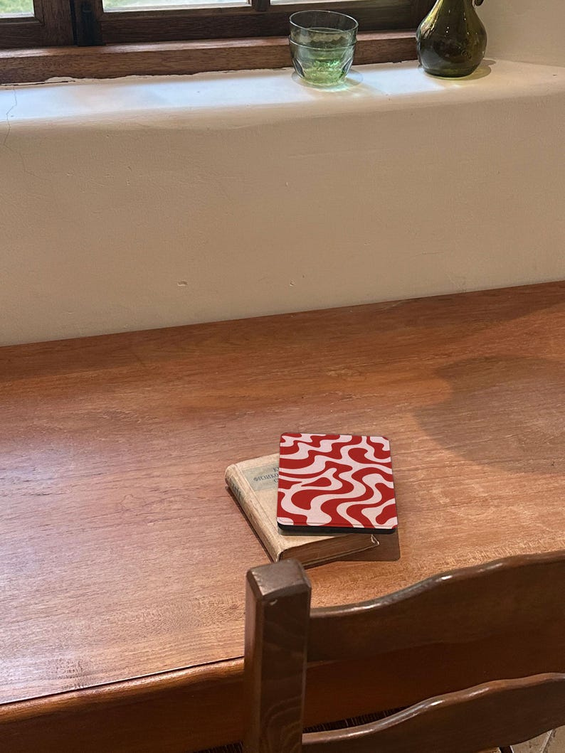Puede incluir: Un posavasos con un patr&oacute;n ondulado rojo y blanco descansa sobre un libro peque&ntilde;o en una mesa de madera. El dise&ntilde;o del posavasos es un patr&oacute;n repetitivo de l&iacute;neas onduladas. Una silla de madera y una ventana con un vaso verde y un jarr&oacute;n est&aacute;n en el fondo.