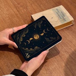 Pode incluir: Uma capa preta para leitor de e-books com um design celestial dourado de fases da lua, estrelas e nuvens. O canto inferior esquerdo exibe as letras douradas AS. A capa tem formato retangular com cantos arredondados e uma borda branca.