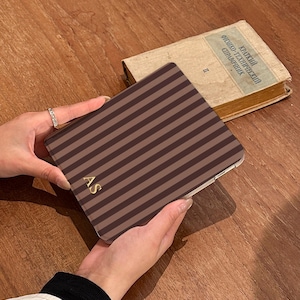 May include: A rectangular electronic tablet case featuring a pattern of vertical dark brown and light brown stripes. The bottom left corner of the case displays the gold-colored initials AS in a serif font. The case has a smooth, flat surface.