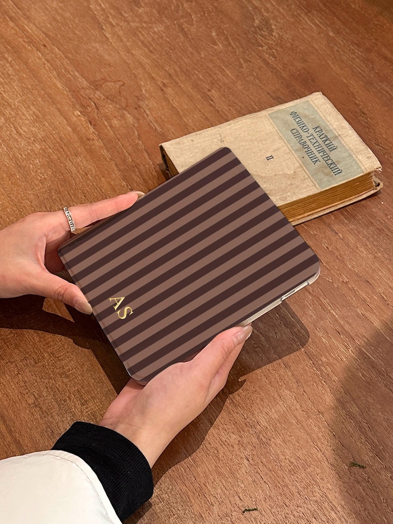 May include: A rectangular electronic tablet case featuring a pattern of vertical dark brown and light brown stripes. The bottom left corner of the case displays the gold-colored initials AS in a serif font. The case has a smooth, flat surface.