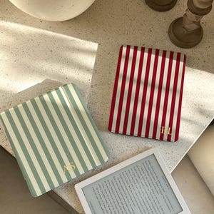 Puede incluir: Tres fundas de libros a rayas rojas y blancas, y verde claro y blanco. Un lector electrónico blanco muestra texto. Iniciales doradas están en las fundas. Las fundas están sobre una superficie clara con una esfera decorativa blanca y dos candelabros.