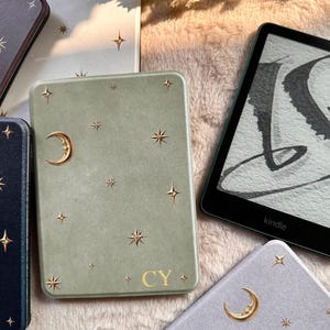 May include: A sage green e-reader case featuring gold-colored celestial motifs including a crescent moon and various star shapes. The bottom center of the case displays the gold-colored letters CY. The case is positioned next to a black Kindle device.