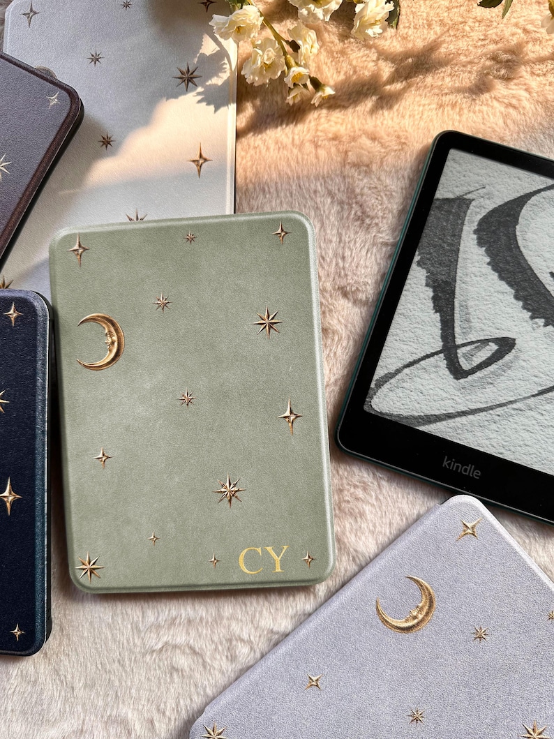 May include: A sage green e-reader case featuring gold-colored celestial motifs including a crescent moon and various star shapes. The bottom center of the case displays the gold-colored letters CY. The case is positioned next to a black Kindle device.
