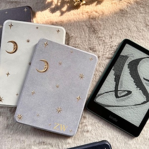 May include: A collection of e-reader cases in white, light gray, and dark blue, each featuring gold foil celestial motifs of crescent moons and stars. The light gray case displays the gold letters ZW at the bottom. A black Kindle e-reader is visible.