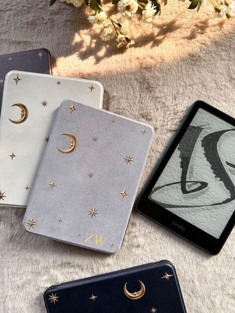 May include: A collection of e-reader cases in white, light gray, and dark blue, each featuring gold foil celestial motifs of crescent moons and stars. The light gray case displays the gold letters ZW at the bottom. A black Kindle e-reader is visible.