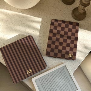 以下が含まれることがあります： 金色のイニシャルが入った3つの茶色のケースと白い電子書籍リーダー。1つのケースはストライプ柄、もう1つは市松模様、3つ目は無地です。電子書籍リーダーには本からのテキストが表示されます。