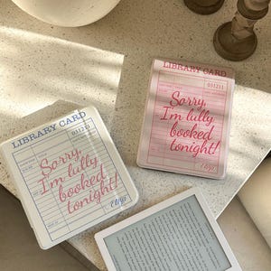 May include: Three square library card-themed items with the text "Sorry, I'm fully booked tonight!" in pink script. One is a white e-reader. The other two are square, one pink and one white, with a library card design.
