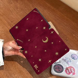 May include: A deep red e-reader case featuring a gold celestial pattern of stars, a crescent moon, and a shooting star. The bottom left corner displays the gold text "BL" flanked by two small stars. The case has a smooth, matte texture.