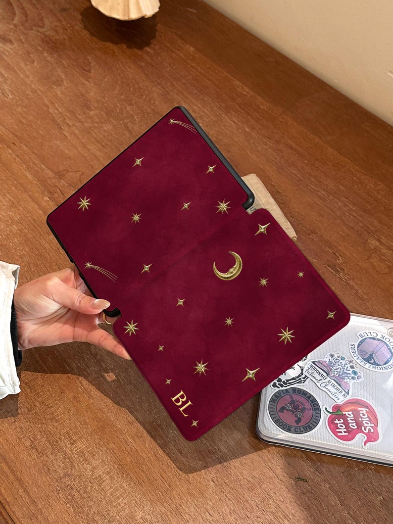 May include: A deep red e-reader case featuring a gold celestial pattern of stars, a crescent moon, and a shooting star. The bottom left corner displays the gold text "BL" flanked by two small stars. The case has a smooth, matte texture.