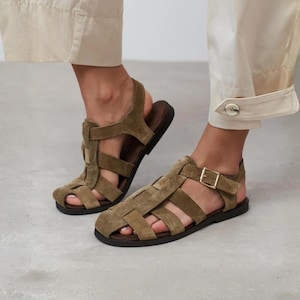 May include: Olive green suede leather fisherman sandals featuring a woven cage design, a closed rounded toe, and an adjustable ankle strap with a gold-toned metal buckle. The sandals have a flat, dark brown sole and a secure back heel strap.