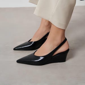 Puede incluir: Zapatos negros de tacón de cuña con punta y correa trasera. Los zapatos tienen un acabado brillante y un tacón de cuña bajo. La correa trasera añade un toque de elegancia. Estos zapatos son un estilo clásico, adecuados para diversas ocasiones.