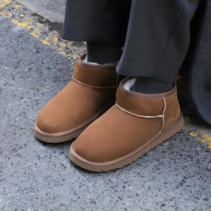 May include: Brown suede ankle boots with a light brown sole. The boots feature a rhinestone trim and white faux fur lining. The boots are worn with dark grey trousers and socks. The boots are suitable for cold weather.