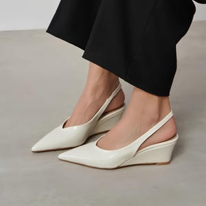 Puede incluir: Zapatos blancos de tacón de cuña con punta. Los zapatos tienen un tacón de cuña bajo y una correa que rodea el tobillo. Los zapatos están hechos de un material brillante.