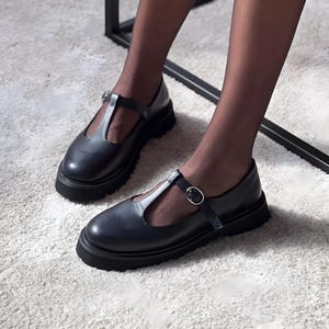 Könnte beinhalten: Schwarze Mary Jane-Schuhe aus Leder mit T-Riemen und Schnallenverschluss. Die Schuhe haben eine klobige Sohle und werden auf einem weißen Teppich gezeigt.