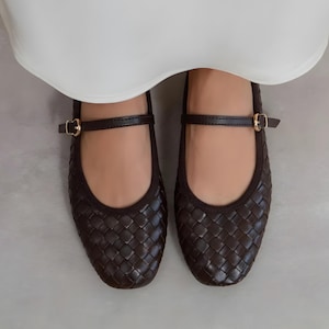 Peut inclure: Une paire de ballerines Mary Jane en cuir tressé marron foncé avec une bride et une boucle dorée. Les chaussures ont un bout arrondi et sont portées avec un vêtement blanc.