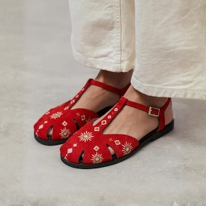 Peut inclure: Sandales en daim rouge avec une bride en T et une boucle dorée réglable. La tige est ornée de motifs brodés blancs et dorés en forme de soleil et de losange, complétée par une semelle plate noire et un style fermé à découpes.