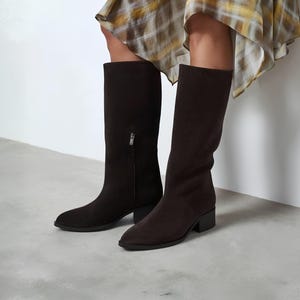 May include: A pair of dark brown knee-high boots with a smooth matte finish, featuring a pointed toe, a low block heel, and a vertical side zipper with a silver-toned metal pull. The boots are worn with a yellow and white plaid skirt.