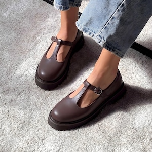 May include: Brown leather T-strap shoes with a buckle closure and a thick, rounded sole. The shoes are worn with blue jeans and are set on a light gray carpet.